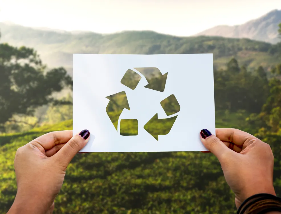 A close-up of two hands holding a Recycling sign in front of a landscape that includes pastureland and mountains.